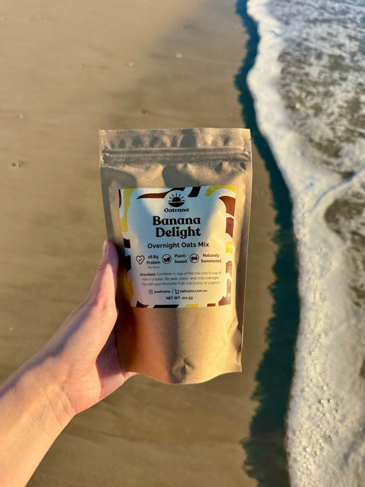 Hand holding a pouch of Oatvana  'Banana Delight' overnight oats mix on a beach.